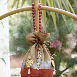 Shimmering tissue silk potli bag featuring an antique gold, silver, and rust color-blocked design with central Kundan stone work and a luxury red beaded handle by The Swamini by Sushmita.