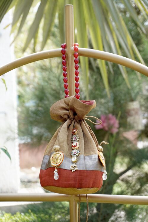 Shimmering tissue silk potli bag featuring an antique gold, silver, and rust color-blocked design with central Kundan stone work and a luxury red beaded handle by The Swamini by Sushmita.