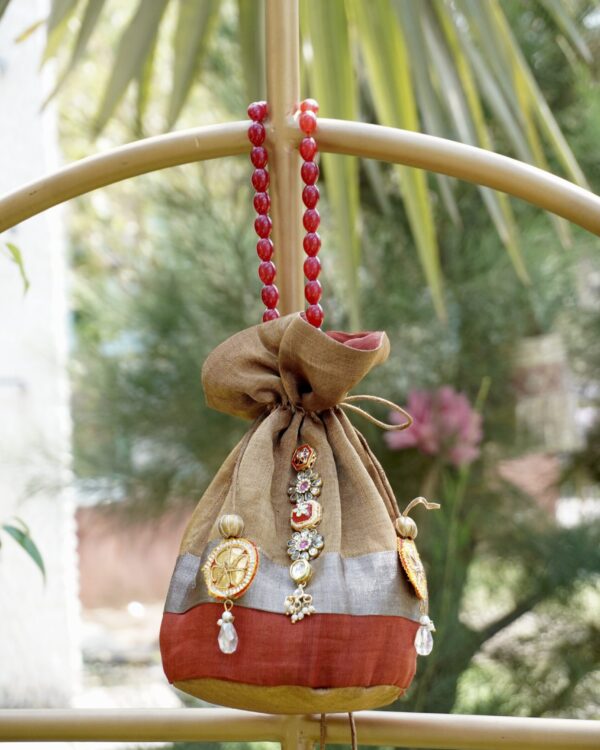 Shimmering tissue silk potli bag featuring an antique gold, silver, and rust color-blocked design with central Kundan stone work and a luxury red beaded handle by The Swamini by Sushmita.