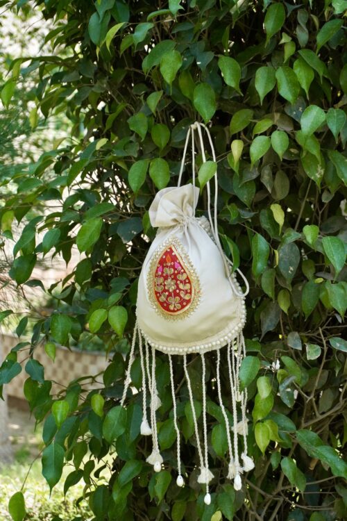 Off-white silk potli bag featuring a vibrant red and gold embroidered paisley motif patch, a pearl border, and long hanging drawstring tassels accented with traditional white cowrie shells by The Swamini by Sushmita.