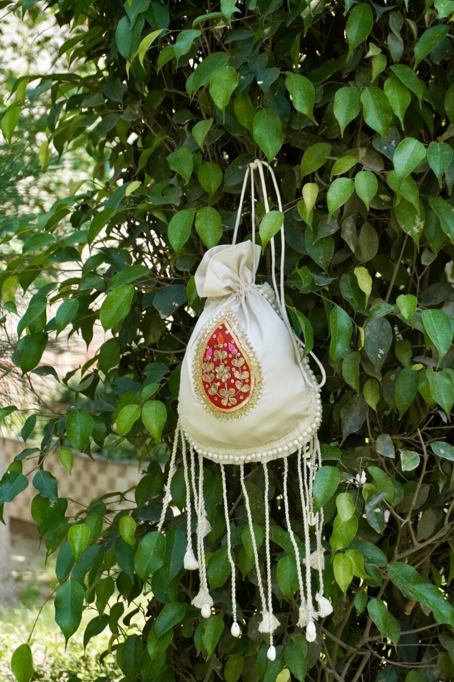 Off-white silk potli bag featuring a vibrant red and gold embroidered paisley motif patch, a pearl border, and long hanging drawstring tassels accented with traditional white cowrie shells by The Swamini by Sushmita.