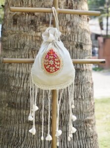 Off-white silk potli bag featuring a vibrant red and gold embroidered paisley motif patch, a pearl border, and long hanging drawstring tassels accented with traditional white cowrie shells by The Swamini by Sushmita.