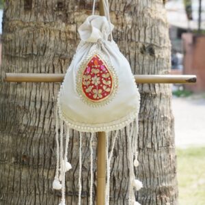 Off-white silk potli bag featuring a vibrant red and gold embroidered paisley motif patch, a pearl border, and long hanging drawstring tassels accented with traditional white cowrie shells by The Swamini by Sushmita.
