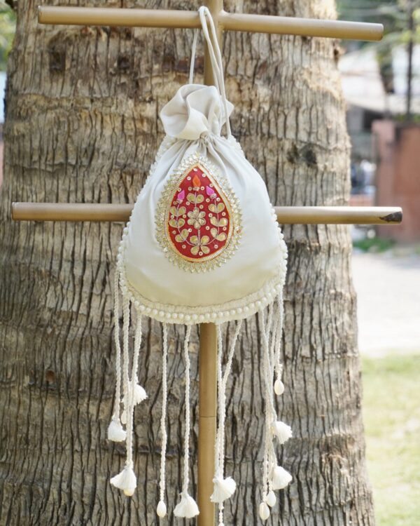 Off-white silk potli bag featuring a vibrant red and gold embroidered paisley motif patch, a pearl border, and long hanging drawstring tassels accented with traditional white cowrie shells by The Swamini by Sushmita.