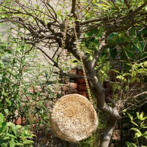 Contemporary circular metallic gold sling bag densely hand-embroidered with shimmering glass bugle beads in a spiral sunburst pattern, hanging from a tree branch by a gold chain strap, by The Swamini by Sushmita.