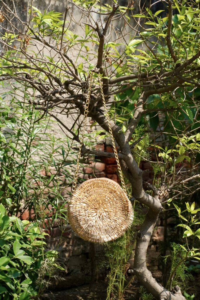 Contemporary circular metallic gold sling bag densely hand-embroidered with shimmering glass bugle beads in a spiral sunburst pattern, hanging from a tree branch by a gold chain strap, by The Swamini by Sushmita.