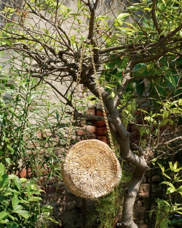 Contemporary circular metallic gold sling bag densely hand-embroidered with shimmering glass bugle beads in a spiral sunburst pattern, hanging from a tree branch by a gold chain strap, by The Swamini by Sushmita.