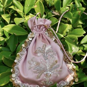 Pastel onion pink satin potli bag featuring central silver and gold sequin floral bouquet embroidery, bordered with pearl lace and hanging gold coins, photographed against green leaves by The Swamini by Sushmita.