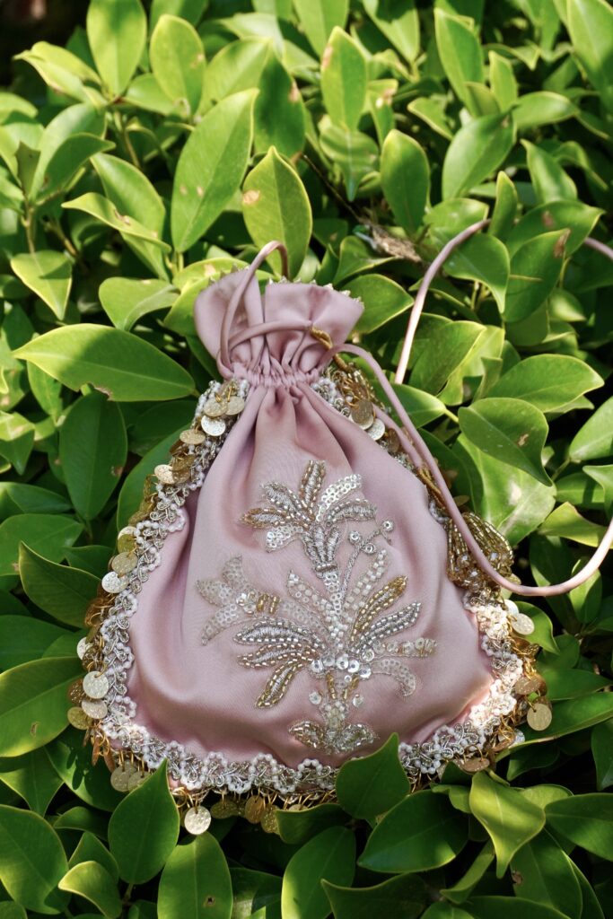 Pastel onion pink satin potli bag featuring central silver and gold sequin floral bouquet embroidery, bordered with pearl lace and hanging gold coins, photographed against green leaves by The Swamini by Sushmita.
