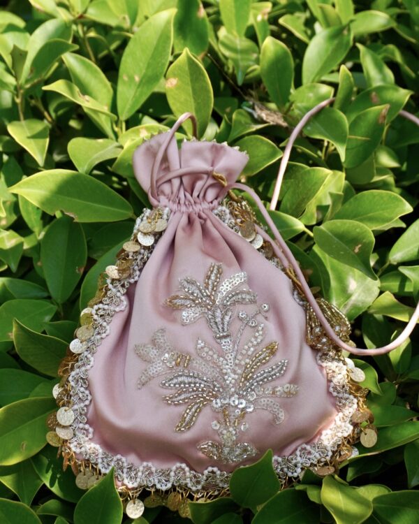 Pastel onion pink satin potli bag featuring central silver and gold sequin floral bouquet embroidery, bordered with pearl lace and hanging gold coins, photographed against green leaves by The Swamini by Sushmita.
