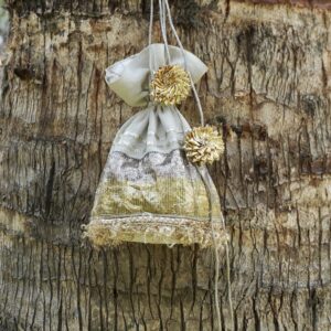 Off-white tissue silk potli bag featuring horizontal shimmering gold and silver panels, fine gold fringe lace at the base, and large pom-pom style gold Gota flower tassels by The Swamini by Sushmita.