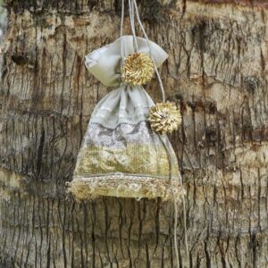 Off-white tissue silk potli bag featuring horizontal shimmering gold and silver panels, fine gold fringe lace at the base, and large pom-pom style gold Gota flower tassels by The Swamini by Sushmita.