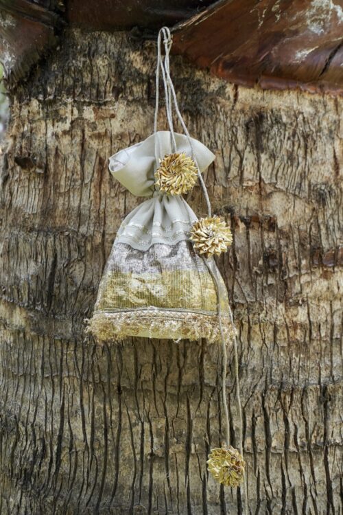 Off-white tissue silk potli bag featuring horizontal shimmering gold and silver panels, fine gold fringe lace at the base, and large pom-pom style gold Gota flower tassels by The Swamini by Sushmita.