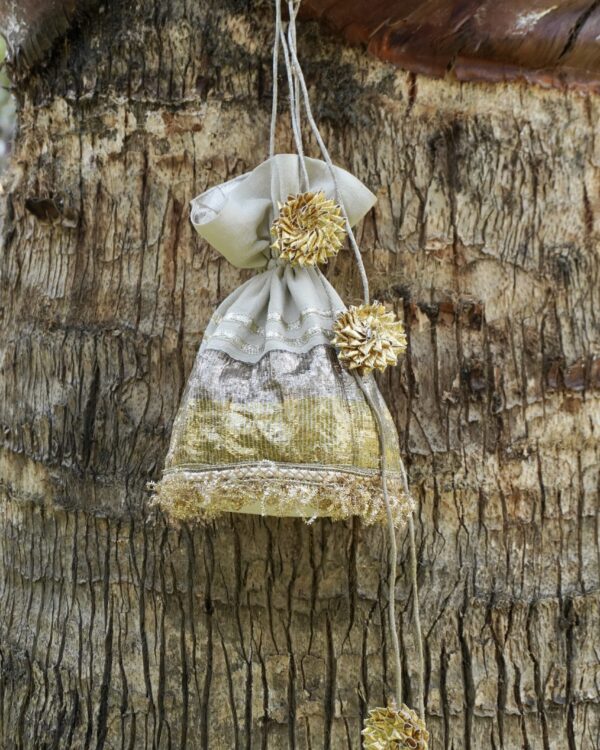 Off-white tissue silk potli bag featuring horizontal shimmering gold and silver panels, fine gold fringe lace at the base, and large pom-pom style gold Gota flower tassels by The Swamini by Sushmita.
