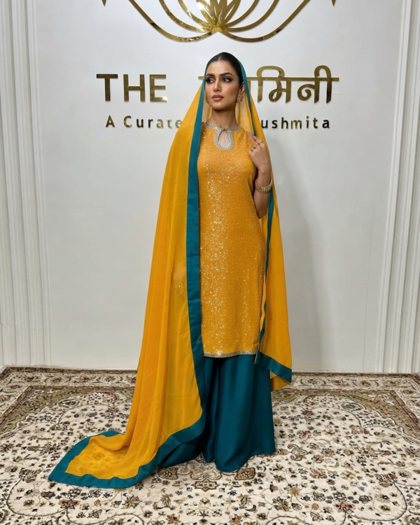 A woman in a mustard-yellow sequin kurta and teal-green palazzo pants with a yellow and green dupatta, posing in front of a branded boutique wall.