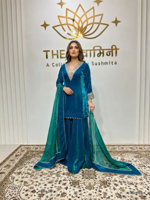 A model in a teal velvet gharara suit with mirrorwork embroidery and a long green dupatta, standing in a branded boutique.