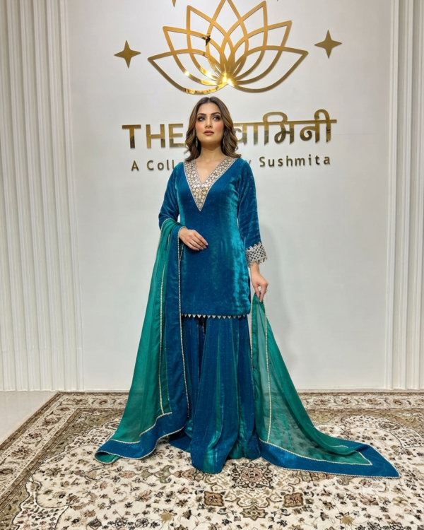 A model in a teal velvet gharara suit with mirrorwork embroidery and a long green dupatta, standing in a branded boutique.