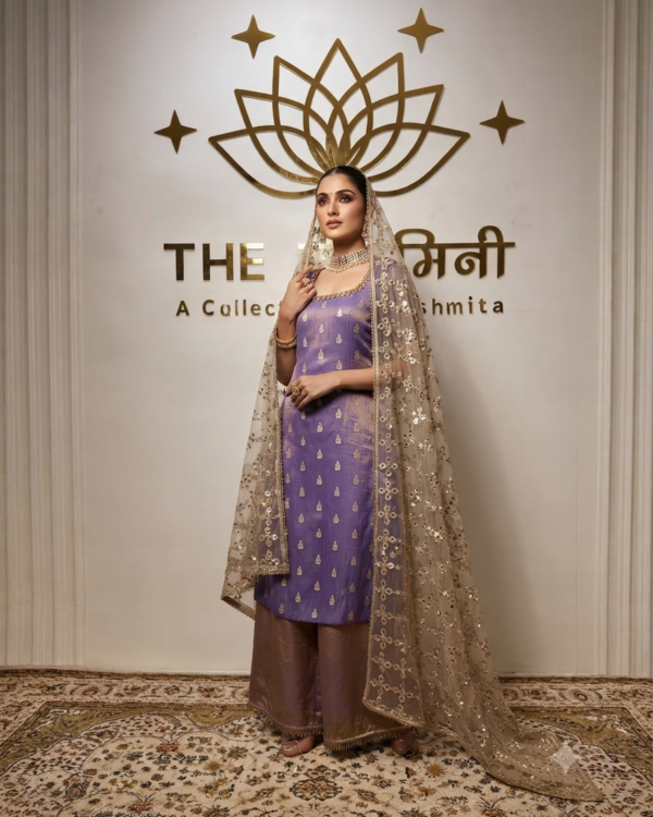 A woman in a lavender-violet Fenty silk suit with gold embroidery and a heavy gold-sequined net dupatta, posing in front of a branded wall.